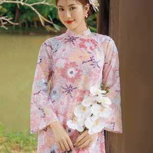 May include: A woman wearing a pink and white floral dress with a white orchid in her hand. She is standing in front of a wooden door.