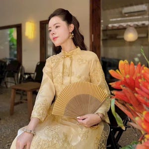 May include: A woman wearing a gold-colored traditional dress with intricate embroidery and a matching fan. The dress features a high collar and long sleeves. The fan is made of light brown paper. The background is a blurred interior with natural light.
