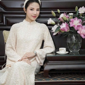 May include: A woman wearing a white sequined dress with a white headband sits on a couch with a vase of pink lilies on a table beside her.