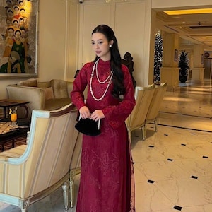 May include: A woman wearing a red dress with a pearl necklace and a black clutch. She is standing in a hotel lobby with a Christmas tree in the background.