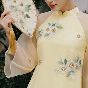 May include: A yellow dress with white floral embroidery and sheer sleeves. The dress has a mandarin collar and a fitted silhouette. The model is holding a white fan with floral embellishments.