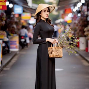 May include: A woman wearing a black dress and a conical straw hat holds a woven basket and a branch with green leaves. She is standing on a street in a city.