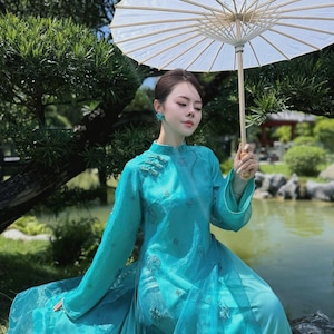 May include: A woman wearing a turquoise dress and holding a white parasol. The dress features long sleeves and embroidered details. The background includes a pond, greenery, and a sunny day.