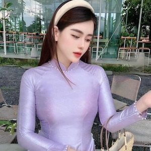 May include: A woman wearing a lavender long-sleeved dress with pearl accents. She is holding a woven straw tote bag with a brown leather handle.