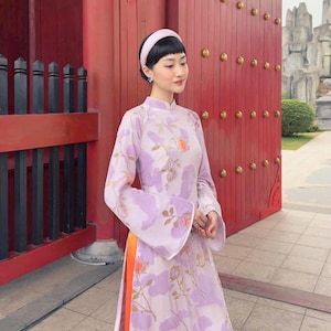 May include: A light purple traditional dress with a floral pattern and long, flared sleeves. The dress features an orange accent down the side and is paired with a matching headband. The background includes a red door and architectural elements.
