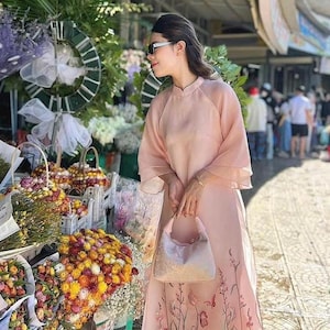 May include: A woman in a pink dress with floral embroidery stands in front of a flower stand. She is wearing sunglasses and holding a small pink handbag.