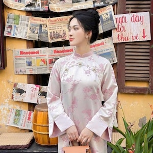 May include: A woman wearing a light pink and white floral patterned dress with long sleeves and a mandarin collar. She is holding a brown handbag. The background features old newspapers and a wooden wall.