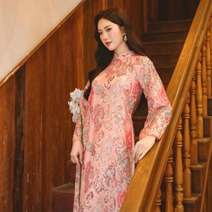 May include: A long, pink dress with an ornate floral pattern and long sleeves. The dress features a mandarin collar and is worn by a person standing on a wooden staircase. The dress has a subtle sheen and is embellished with silver accents.
