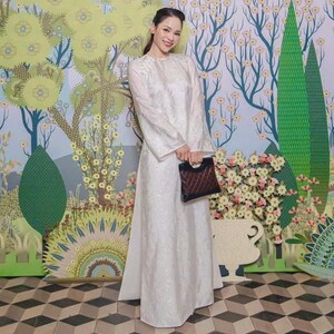 May include: A woman wearing a white, long-sleeved dress with a floral pattern, holding a dark brown quilted handbag. The background features a colorful mural with floral and tree designs, and a black and white checkered floor.