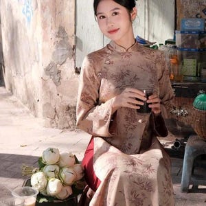 May include: A woman in a beige floral dress with a high collar sits on a stool and holds a glass of dark liquid. She is in front of a building with a weathered wall. A bouquet of white lotus flowers sits on a green stool in front of her.