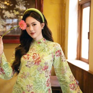 May include: A light green dress with a floral pattern featuring red and pink flowers. The dress has long sleeves and a mandarin collar. A green headband and a pink flower are also visible.
