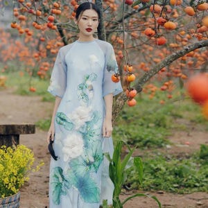 May include: A woman wearing a light blue dress with a white floral print and green pants stands in front of a tree with orange fruit.