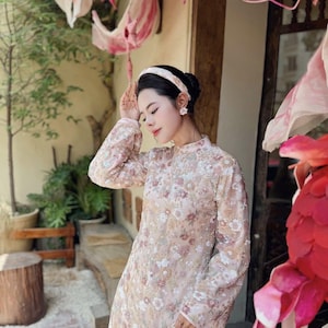 May include: A light pink, long-sleeved dress with a floral pattern. The dress has a mandarin collar and is adorned with delicate embroidery. The woman is wearing a matching headband and earrings. The background features pink floral decorations and greenery.