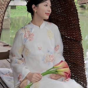 May include: A woman wearing a white, long-sleeved dress with floral embroidery, holding a bouquet of calla lilies. The dress features a mandarin collar and is adorned with pink and yellow floral patterns. The background shows a serene waterscape.