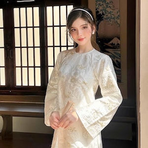 May include: A woman wearing a white traditional dress with gold embroidery and a white headband. She is holding a wooden fan in her hands.