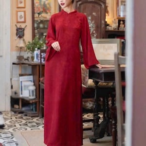 May include: A long, flowing red dress with long sleeves and a mandarin collar. The dress has a subtle floral pattern and is paired with matching red pants. The woman is standing in a room with antique furniture and decor.