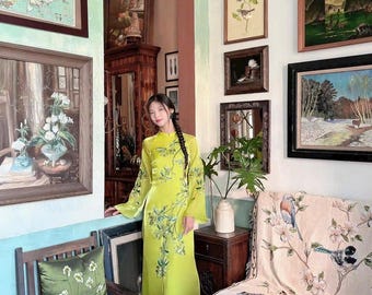 May include: A bright lime green long-sleeved tunic dress featuring a high collar and delicate green leaf embroidery patterns. The garment is floor-length with a straight silhouette and long, flowing sleeves, worn over dark teal trousers.