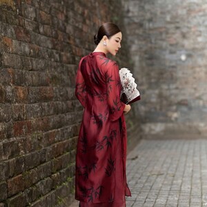 May include: A woman in a red dress with a black bamboo print stands in front of a brick wall. She is holding a white and black hand fan.