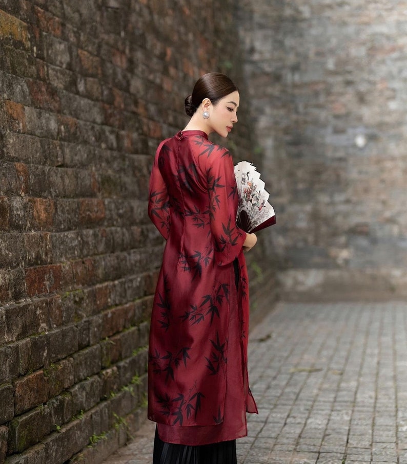 May include: A woman in a red dress with a black bamboo print stands in front of a brick wall. She is holding a white and black hand fan.