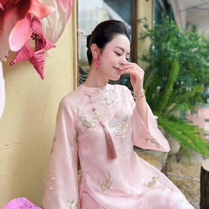 May include: A light pink, long-sleeved dress with floral embroidery and a tassel necklace. The dress features wide sleeves and is adorned with delicate embroidered flowers and fish. The woman is wearing pink earrings.