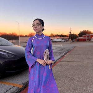 May include: A woman wearing a purple and pink patterned dress with a white collar and a white flower design. She is holding a white fan and is standing in front of a car.