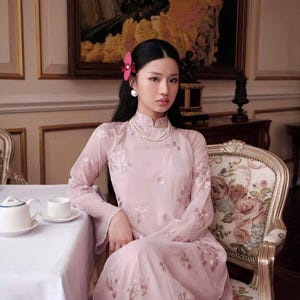 May include: A woman wearing a pink, floral embroidered dress with a pearl necklace and a red flower in her hair. She is sitting in a chair in a room with a white tablecloth and a painting on the wall.