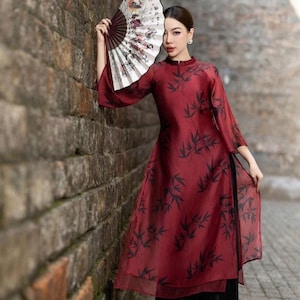 May include: A woman wearing a red dress with a black bamboo print and a black skirt. She is holding a white and black hand fan in front of her face. The dress has a high neck and long sleeves. The woman is standing in front of a brick wall.