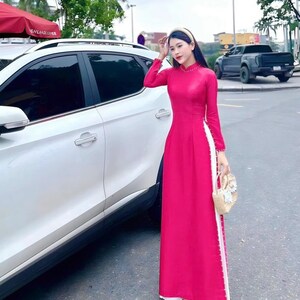 May include: A woman wearing a pink dress with white trim and a white headband. She is standing in front of a white car and holding a straw bag.