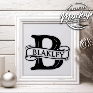 Könnte beinhalten: Gerahmter Druck mit einem schwarzen Monogramm-Buchstaben "B" und dem Namen "Blakley" auf einem Banner. Der Druck befindet sich in einem weißen Rahmen, vor einem weißen Holzgrund. Das Bild enthält eine silberne Kugel und einen Tannenzapfen.