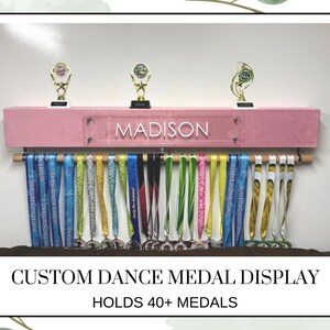 May include: A pink custom dance medal display with the name "MADISON" in white lettering. The display holds over 40 medals and features three gold trophies on top. Colorful ribbons hang below, showcasing dance medals.
