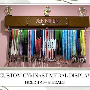 May include: A custom gymnast medal display with a wooden shelf and a rod for hanging medals. The display features a nameplate that reads "JENNIFER" and holds over 40 medals. Gold trophies sit on top of the shelf. The medals have colorful ribbons.