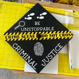 Op de afbeelding: Zwarte afstudeerhoed met een geel en zwart politielint met de tekst "Crime Scene Do Not Cross". De hoed heeft ook een vingerafdruk, een vergrootglas en de tekst "Be Unstoppable Criminal Justice".