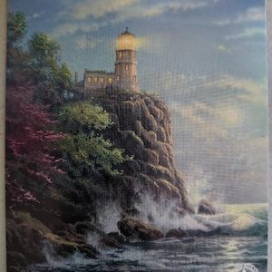 May include: A rectangular print depicting a lighthouse perched on a dark, craggy cliffside at twilight. The lighthouse lantern glows with a warm yellow light. Below, ocean waves crash against the rocks. The text Thomas Kinkade Painter of Light is visible.
