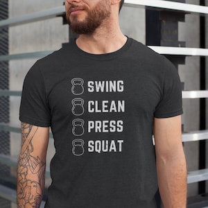 Op de afbeelding: Een donker grijs t-shirt met een witte afbeelding van een kettlebell en de tekst "Swing, Clean, Press, Squat".