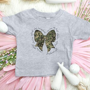 Peut inclure: Un t-shirt gris &agrave; manches courtes avec un n&oelig;ud camouflage et le texte "A hero has my heart, a hero has my heart, a hero has my heart, a hero has my heart".