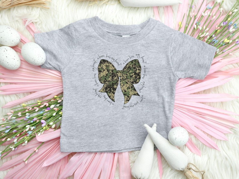 Peut inclure: Un t-shirt gris &agrave; manches courtes avec un n&oelig;ud camouflage et le texte "A hero has my heart, a hero has my heart, a hero has my heart, a hero has my heart".