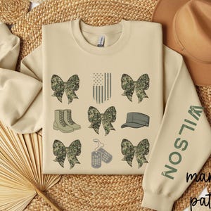 May include: Sand-colored sweatshirt with a marine pattern design. The design includes camouflage bows, boots, a cap, dog tags, and an American flag. The sleeve features the name "WILSON" in green. The words "marine pattern" are printed at the bottom.