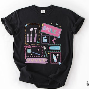 Op de afbeelding: Een zwart t-shirt met een kleurrijke grafische print van tandheelkundige instrumenten, waaronder een spiegel, tandenborstel en tandpastatube. Het ontwerp bevat de tekst "SMILE :)" in een roze tekstballon, omringd door kleine hartjes en kaders.
