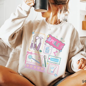 Op de afbeelding: Een zandkleurige sweater met ronde hals en een kleurrijke grafische print van tandheelkundige instrumenten, waaronder een spiegel, tandenborstel, tandpasta en flosdraad, met de tekst "SMILE :)" in een roze tekstballon en kleine hartjes.