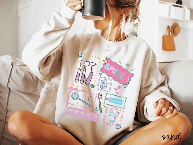 Op de afbeelding: Een zandkleurige sweater met ronde hals en een kleurrijke grafische print van tandheelkundige instrumenten, waaronder een spiegel, tandenborstel, tandpasta en flosdraad, met de tekst "SMILE :)" in een roze tekstballon en kleine hartjes.