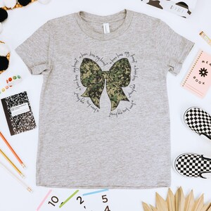 Peut inclure: Un t-shirt gris avec un n&oelig;ud camouflage et le texte "J'ai un h&eacute;ros dans mon c&oelig;ur, j'aime tellement ce gar&ccedil;on, un h&eacute;ros a mon c&oelig;ur, j'aime tellement ce gar&ccedil;on".
