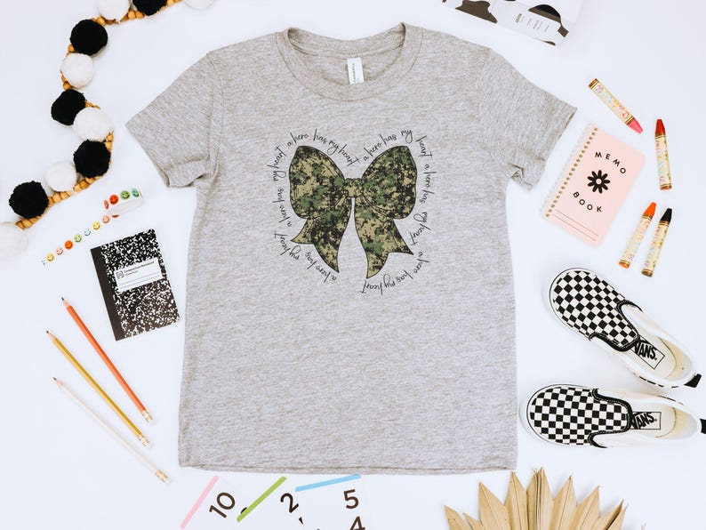 Peut inclure: Un t-shirt gris avec un n&oelig;ud camouflage et le texte "J'ai un h&eacute;ros dans mon c&oelig;ur, j'aime tellement ce gar&ccedil;on, un h&eacute;ros a mon c&oelig;ur, j'aime tellement ce gar&ccedil;on".