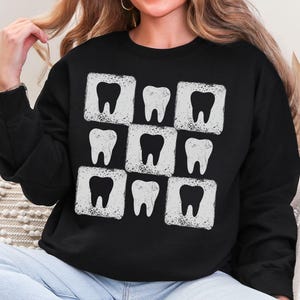 K&ouml;nnte beinhalten: Schwarzes Sweatshirt mit einem sich wiederholenden Muster aus wei&szlig;en Z&auml;hnen in wei&szlig;en quadratischen Rahmen. Die Z&auml;hne haben ein verwittertes, Vintage-Aussehen. Das Sweatshirt ist ein Rundhals-Modell und aus weichem Material.