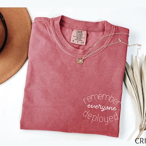 May include: Crimson-colored t-shirt with the words "remember everyone deployed" in white script. A gold necklace with a pendant rests on the shirt. A brown hat and dried plant are also in the image.