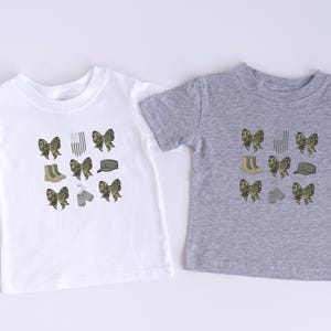 May include: Two t-shirts with a camouflage pattern. One is white and the other is gray. Both shirts feature a design of bows, boots, hats, and dog tags.