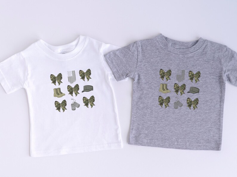 May include: Two t-shirts with a camouflage pattern. One is white and the other is gray. Both shirts feature a design of bows, boots, hats, and dog tags.