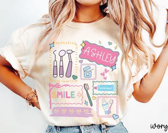 May include: An ivory t-shirt featuring a colorful graphic print of dental tools, including a mirror, probe, and scaler, alongside a toothbrush, toothpaste, and dental floss. The design includes the name ASHLEY and the word SMILE with hearts.