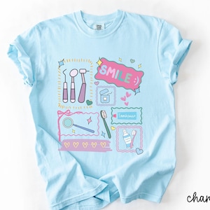 Op de afbeelding: Een lichtblauw t-shirt met een grafische print van tandheelkundige instrumenten, waaronder een spiegel, sonde en scaler, naast een tube tandpasta, een tandenborstel en flosdraad. Het ontwerp bevat de tekst "SMILE :)" en "Toothpaste".