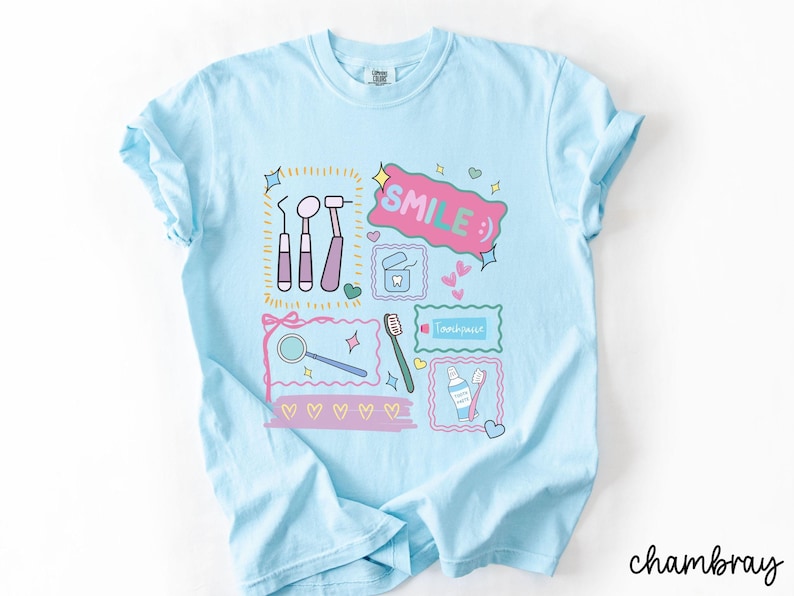 Op de afbeelding: Een lichtblauw t-shirt met een grafische print van tandheelkundige instrumenten, waaronder een spiegel, sonde en scaler, naast een tube tandpasta, een tandenborstel en flosdraad. Het ontwerp bevat de tekst "SMILE :)" en "Toothpaste".