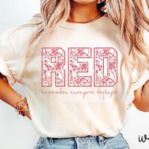 May include: Ivory-colored t-shirt with the word "RED" in a distressed, floral-filled font. Below, the text reads "remember everyone deployed" in a script font. The shirt is a casual style.